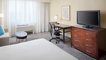 Ett hotellrum med en säng, ett skrivbord, en stol, en tv, en byrå och ett fönster med gardiner.