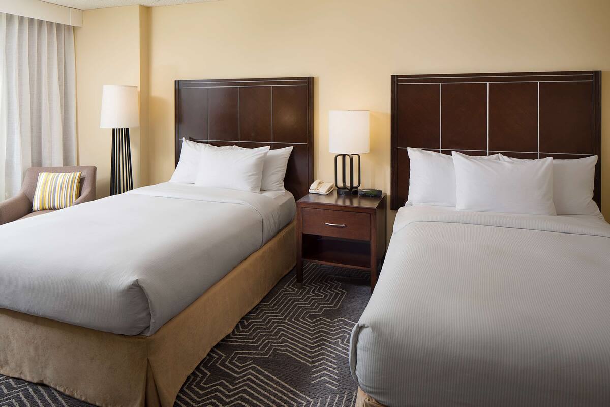 A hotel room with two beds, a nightstand with a lamp, a telephone, and a chair with a striped pillow.