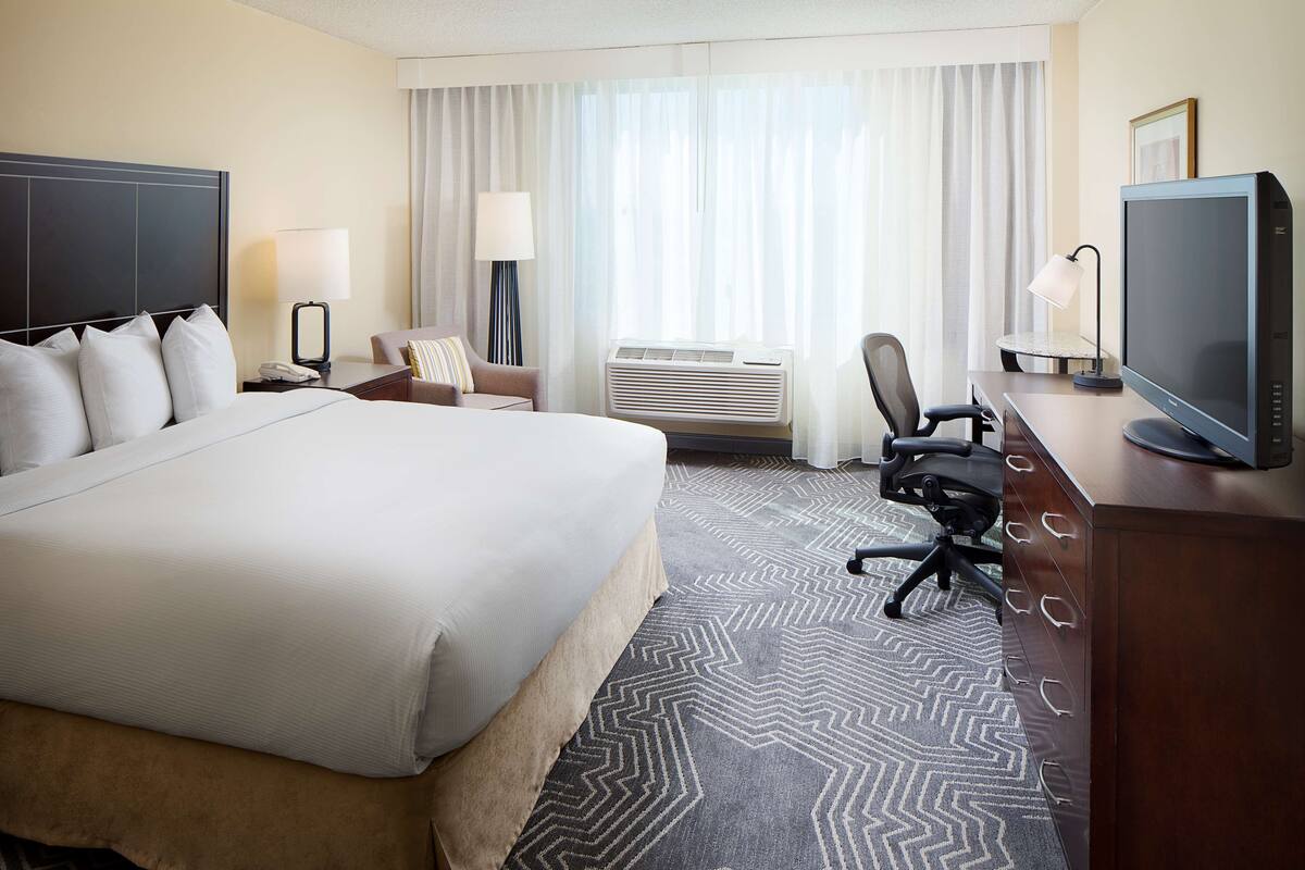 A hotel room with a large bed, a desk with a computer, a chair, an air conditioning unit, and a television.