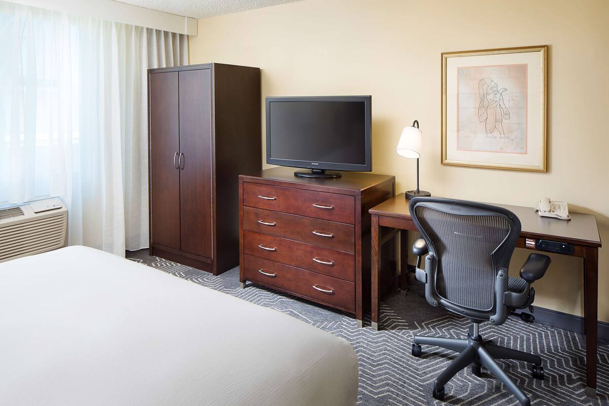 A hotel room with a bed, a desk with a computer, a television, a wardrobe, and a framed picture on the wall.