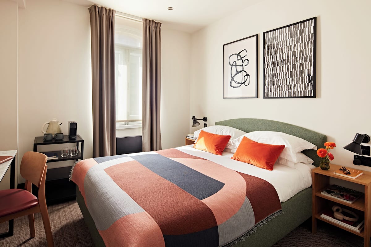 A hotel room with a bed, a desk, a chair, a nightstand, and a window with curtains.