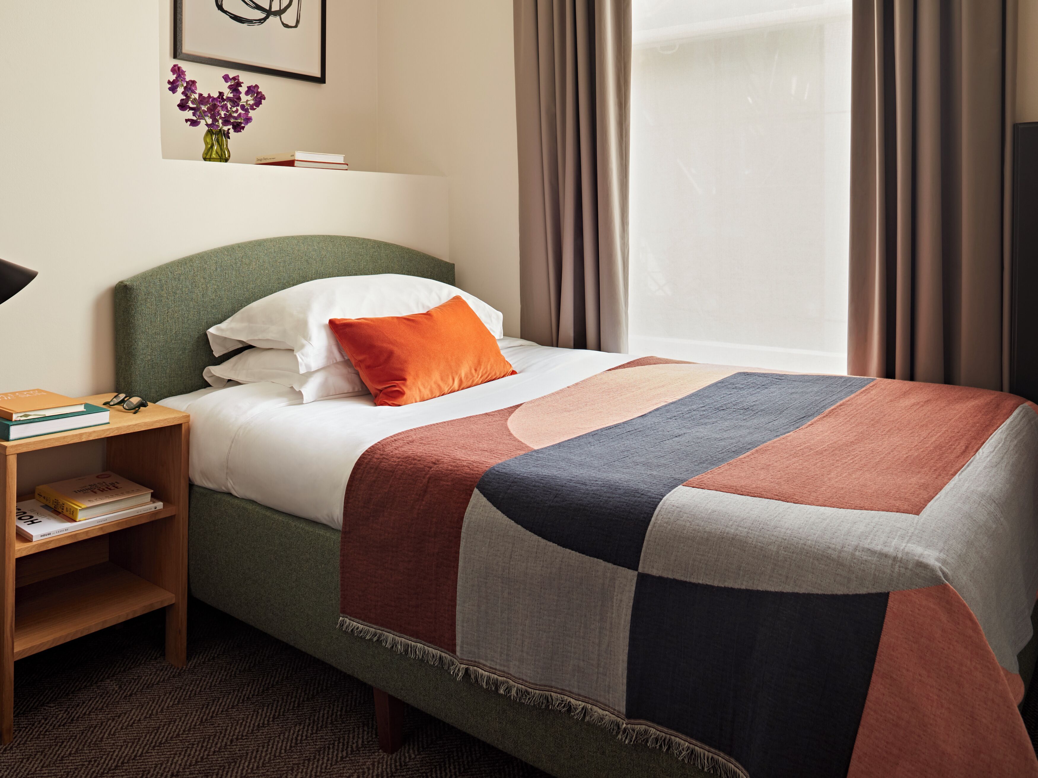 A neatly made bed with a patchwork bedspread, a bedside table with books, a small vase with flowers, and a framed artwork on the wall.
