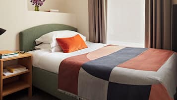 A neatly made bed with a patchwork bedspread, a bedside table with books, a small vase with flowers, and a framed artwork on the wall.
