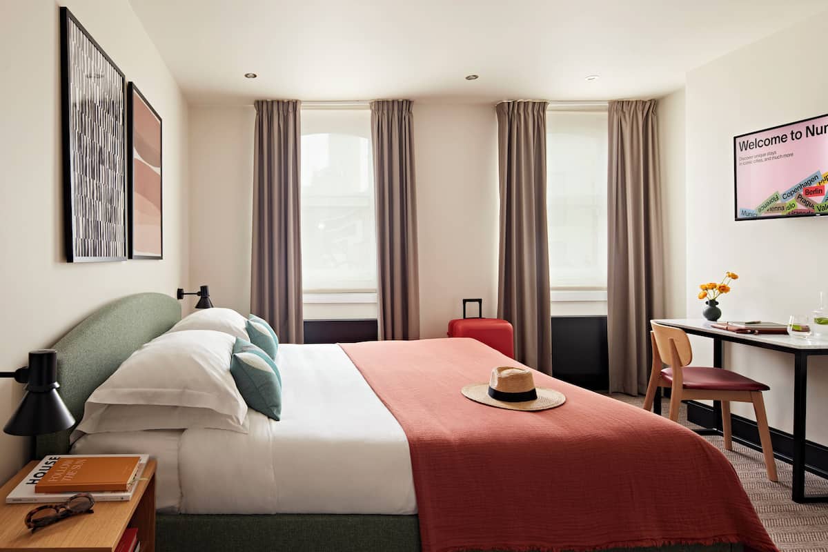 A hotel room with a bed, a desk, a chair, a suitcase, and a framed welcome sign.