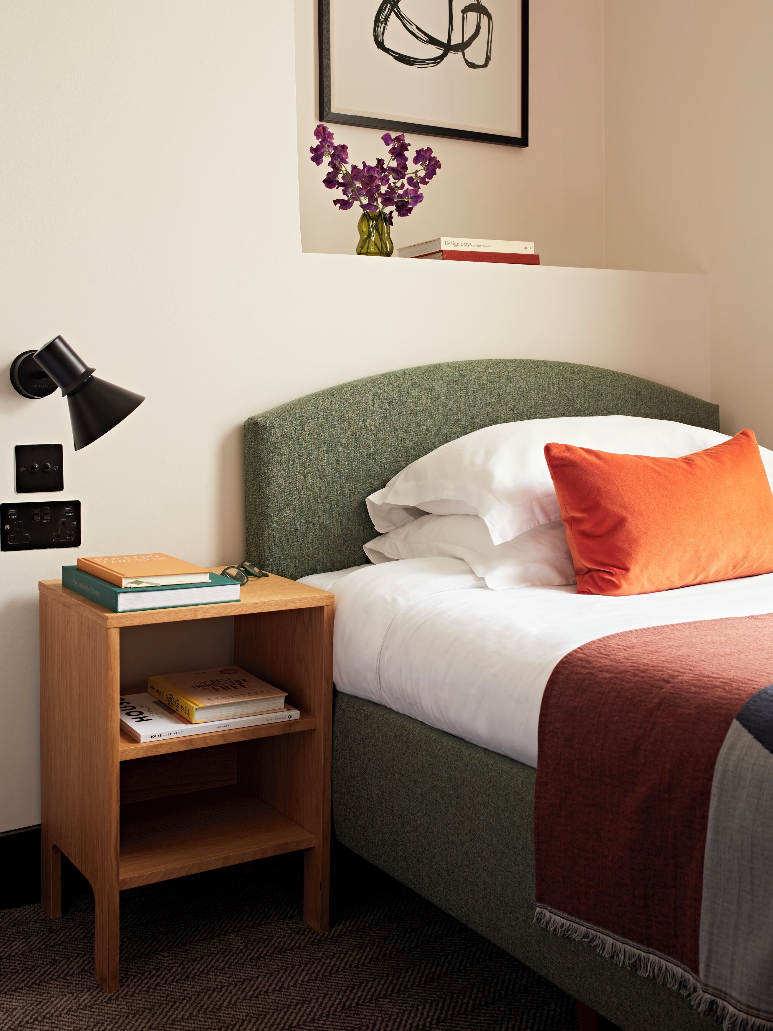 A neatly arranged bedroom with a green upholstered headboard, white bedding, an orange pillow, and a small wooden nightstand with books.