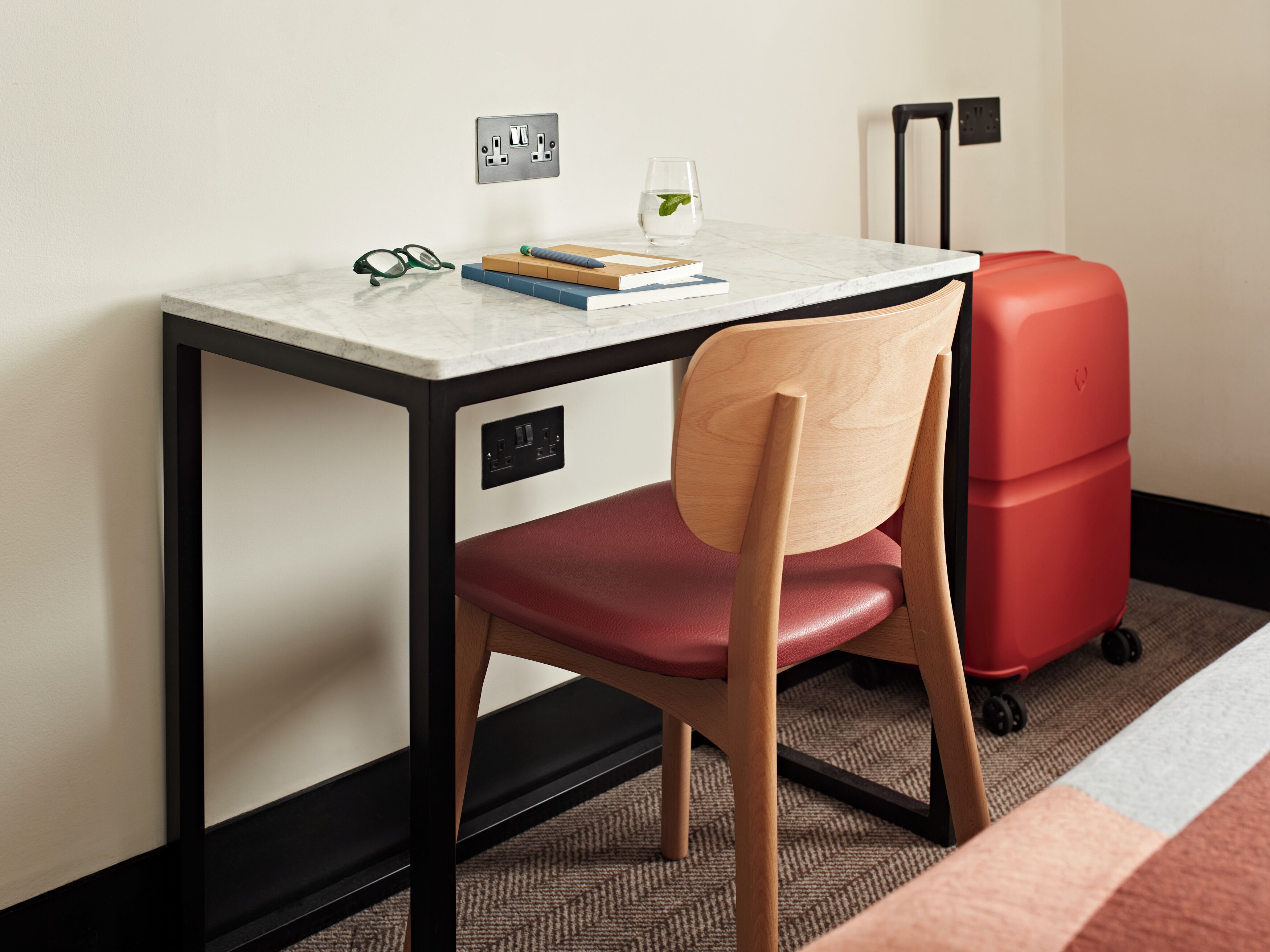 A compact workspace with a desk, chair, and a red suitcase.