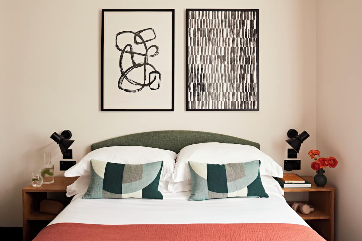 A neatly arranged hotel room with a bed, two pillows, a nightstand with a lamp, a vase with flowers, and two framed artworks on the wall.