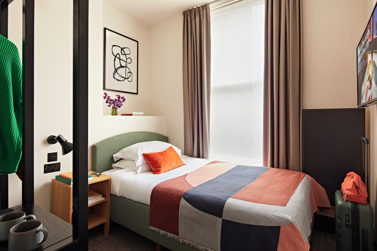 A neatly made bed with a colorful quilt, a bedside lamp, a small nightstand, a green sweater hanging, and a suitcase on the floor.