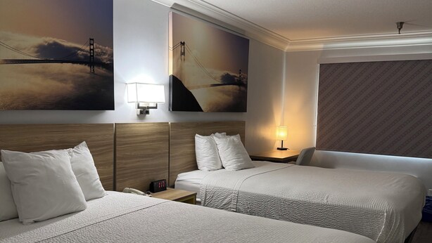 A hotel room with two beds, a nightstand, a lamp, and a painting of a bridge.