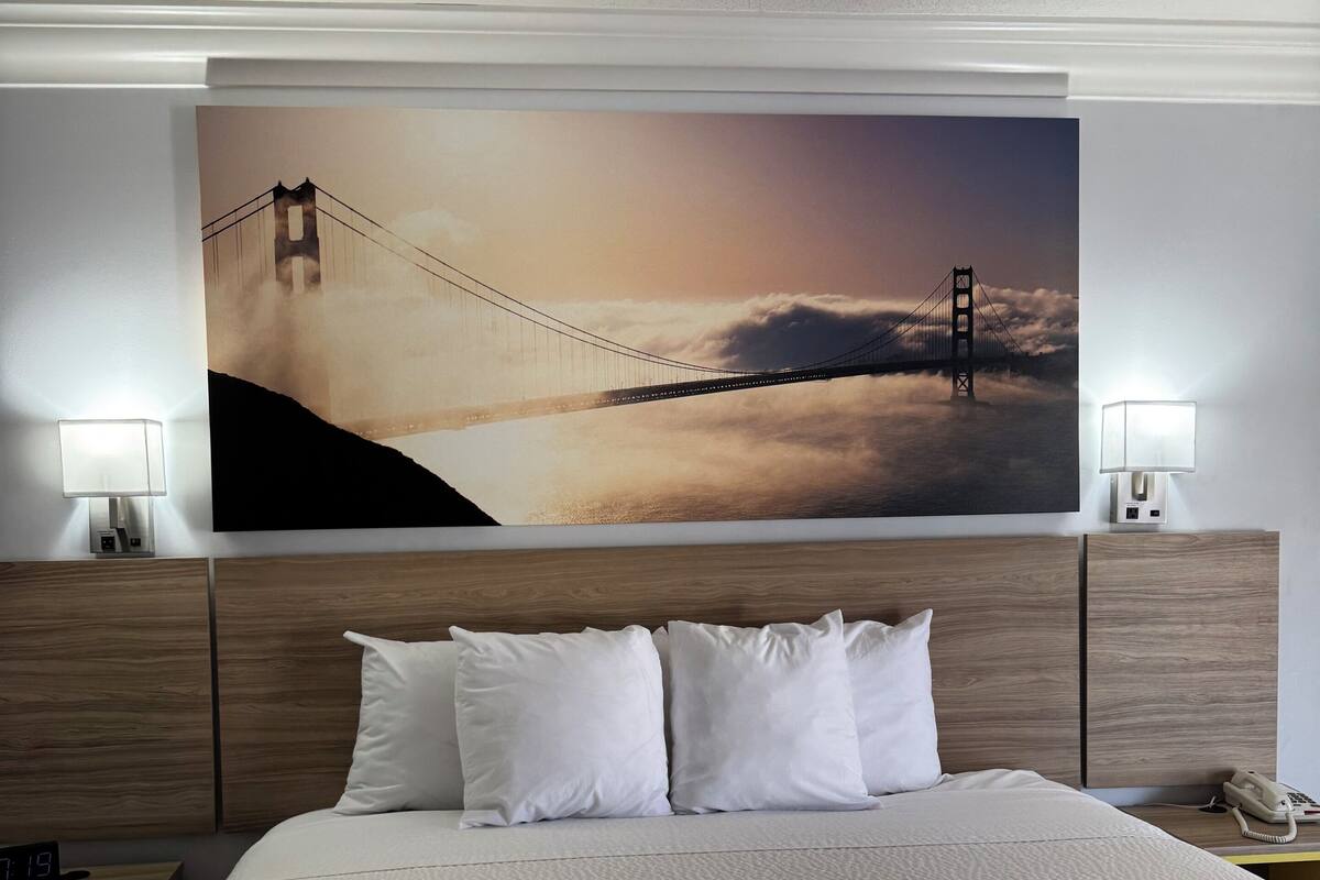 A hotel room with a large bed, two bedside lamps, a nightstand with a phone, and a wall-mounted painting of the Golden Gate Bridge.