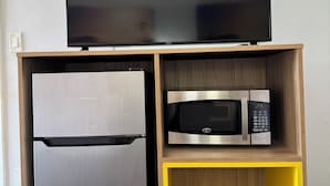 A compact kitchen with a stainless steel refrigerator, a microwave, and a flat-screen TV on a wooden shelf.