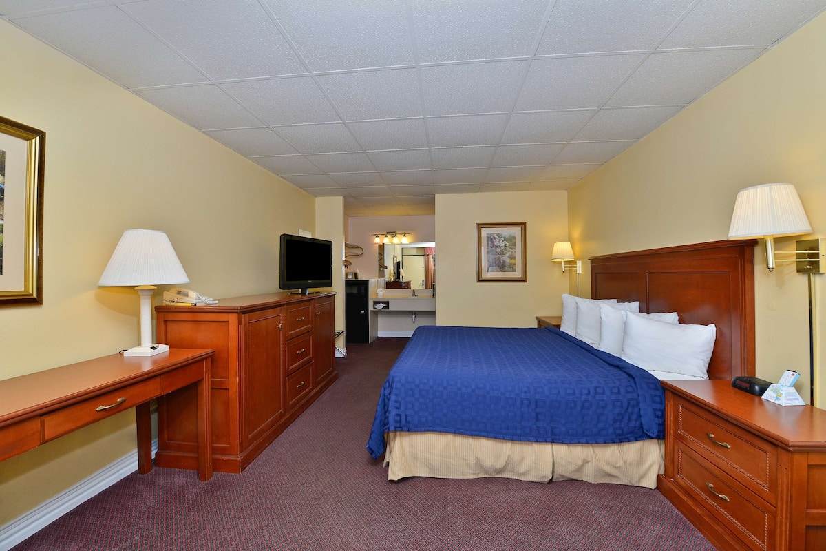 A hotel room with a bed, a desk with a lamp, a dresser, a television, and a bathroom visible in the background.