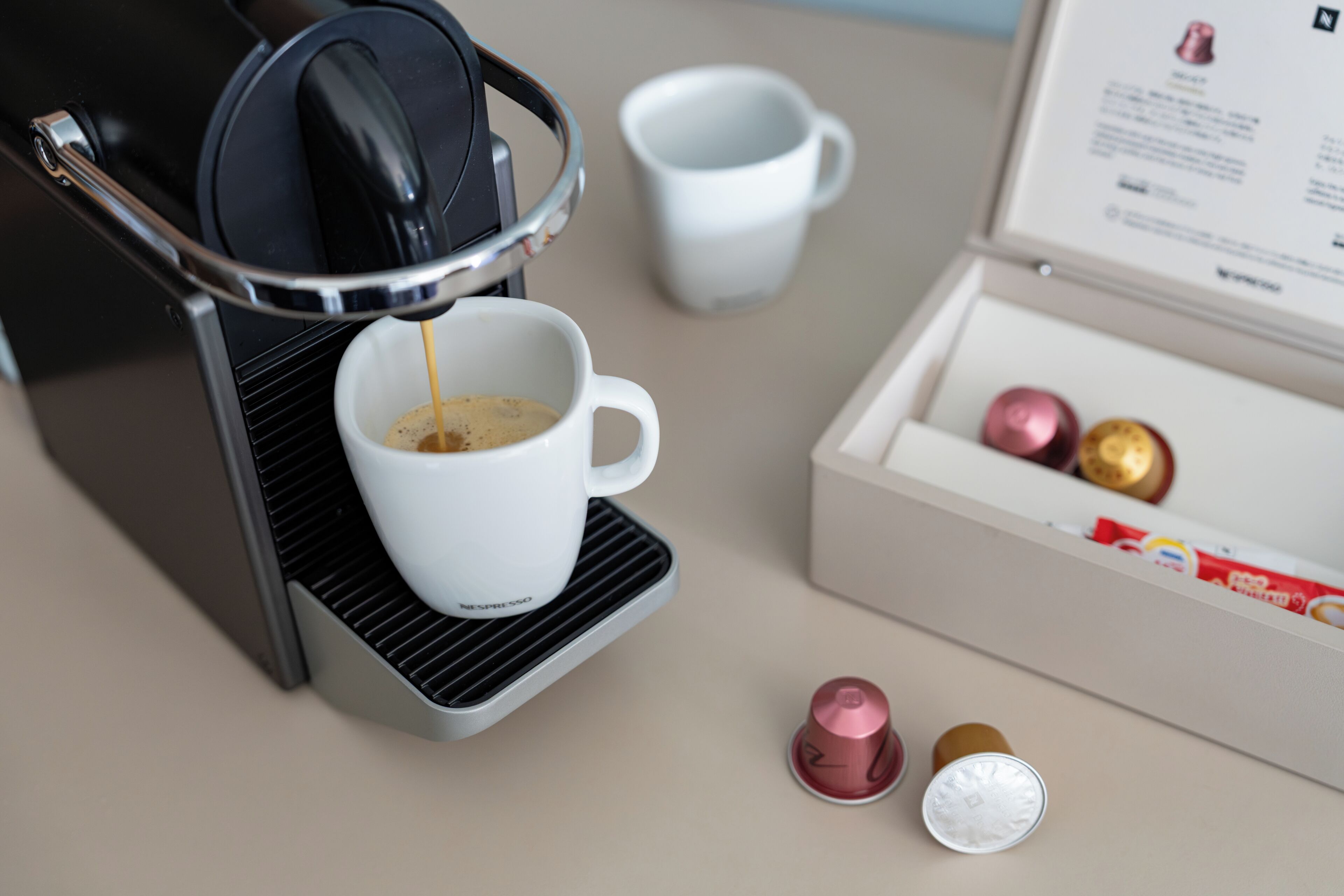 Una cafetera sirviendo café en una taza, una caja de cápsulas de café y una taza blanca sobre una mesa.