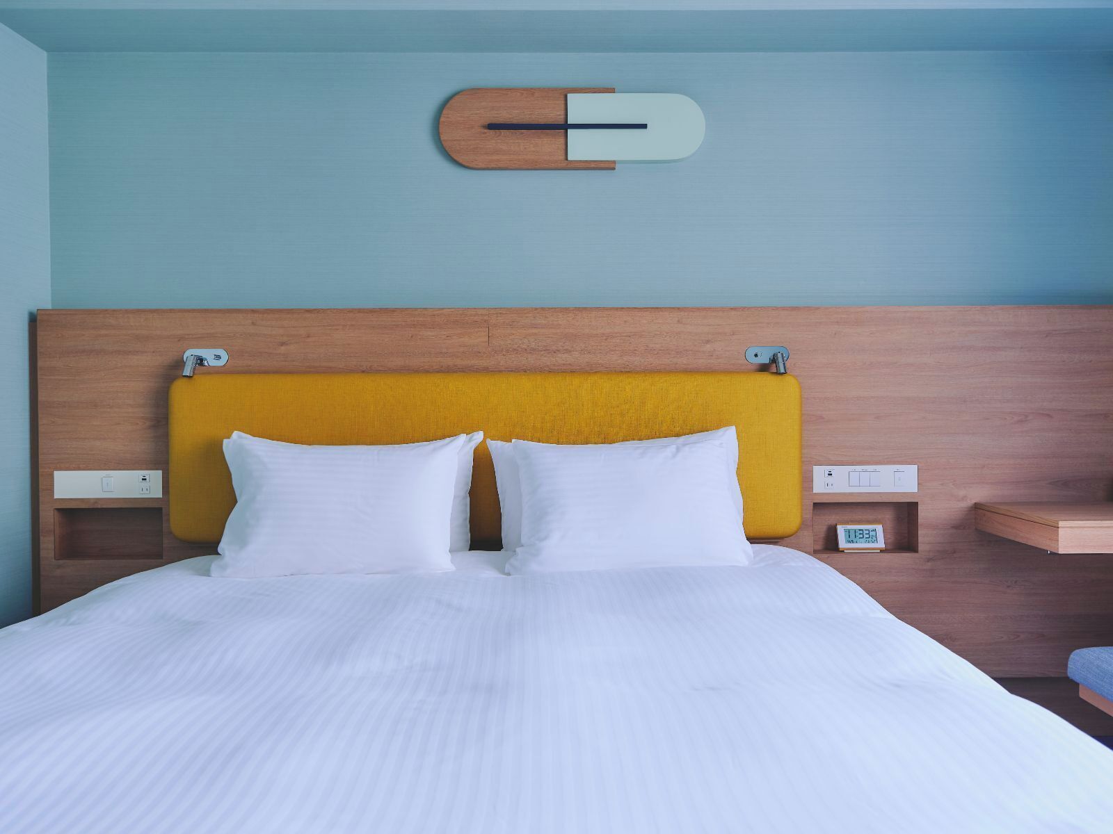 A hotel room with a wooden headboard, a bed with white bedding, and a small bedside table with a clock.