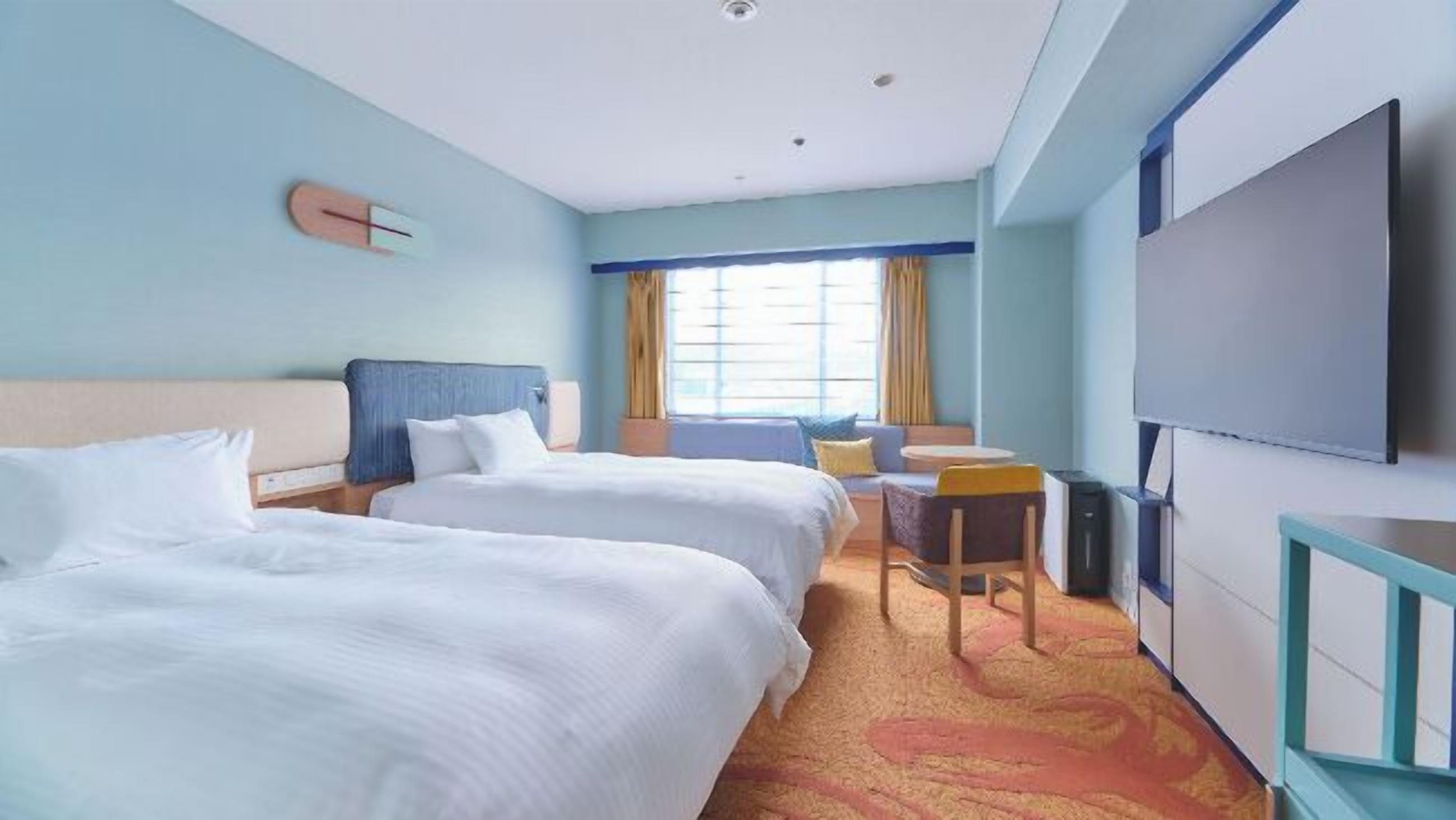 Quarto de hotel com duas camas, uma mesa, uma cadeira, uma televisão e uma janela com cortinas.