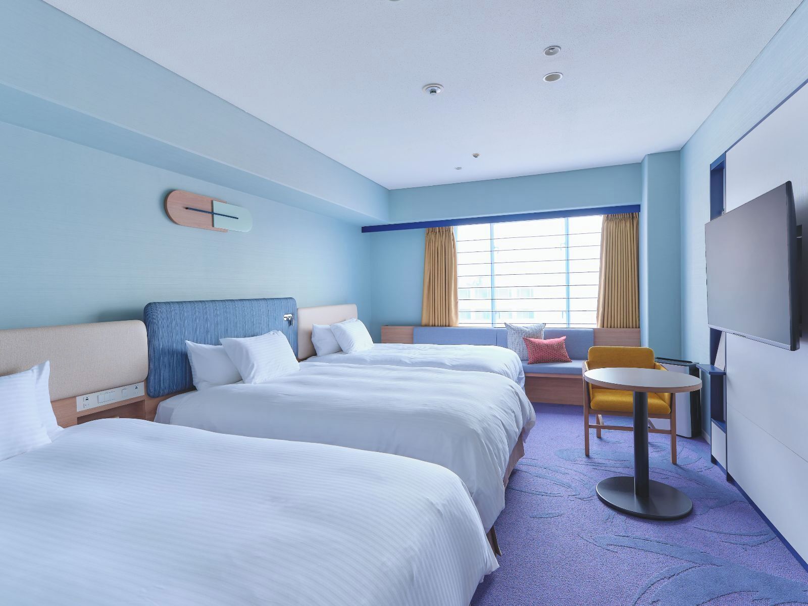 A hotel room with two beds, a small table, a chair, a TV, and a window with curtains.