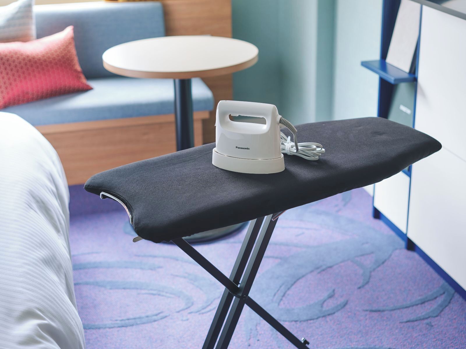 A neatly made bed with white bedding, a black ironing board with a white iron, and a round table with a white top in the background.