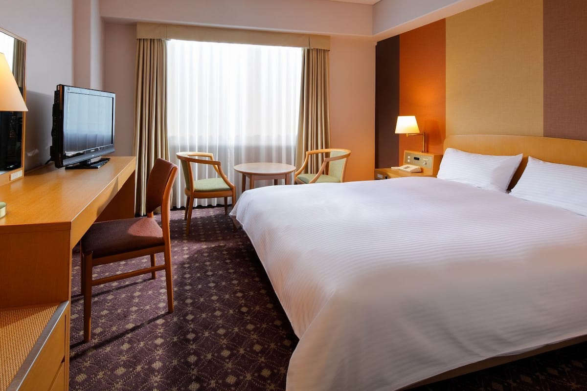 Une chambre d’hôtel comprenant un lit, un bureau avec une télévision, deux chaises, une petite table et une table de chevet avec une lampe.
