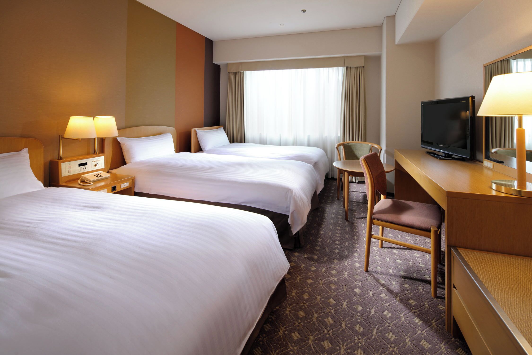 A hotel room with two beds, a desk with a television, a chair, and a desk lamp.