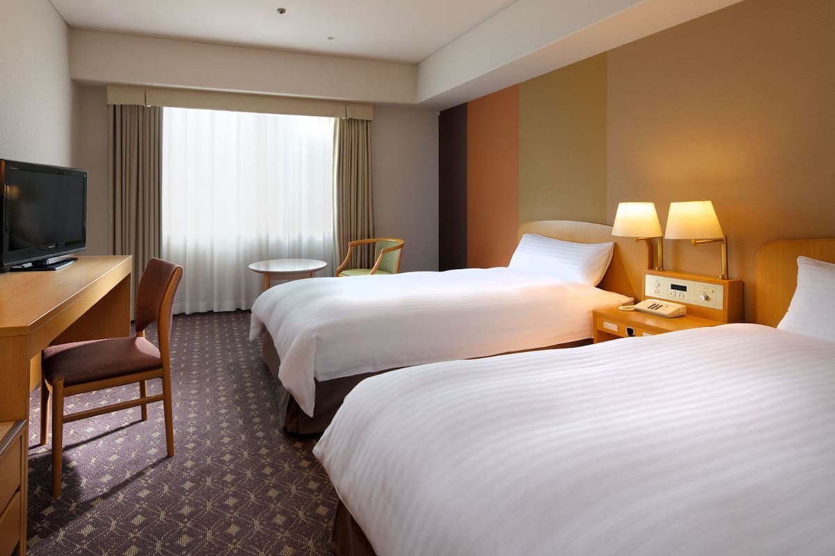 Une chambre d’hôtel avec deux lits, un bureau avec une télévision, une chaise, une petite table et une fenêtre avec des rideaux.