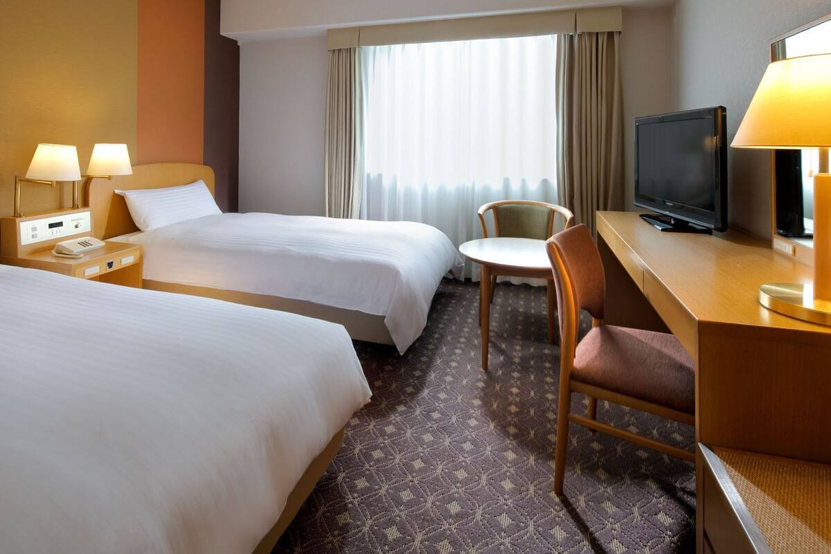 Une chambre d’hôtel avec deux lits, un bureau avec une télévision, une chaise et une fenêtre avec des rideaux.