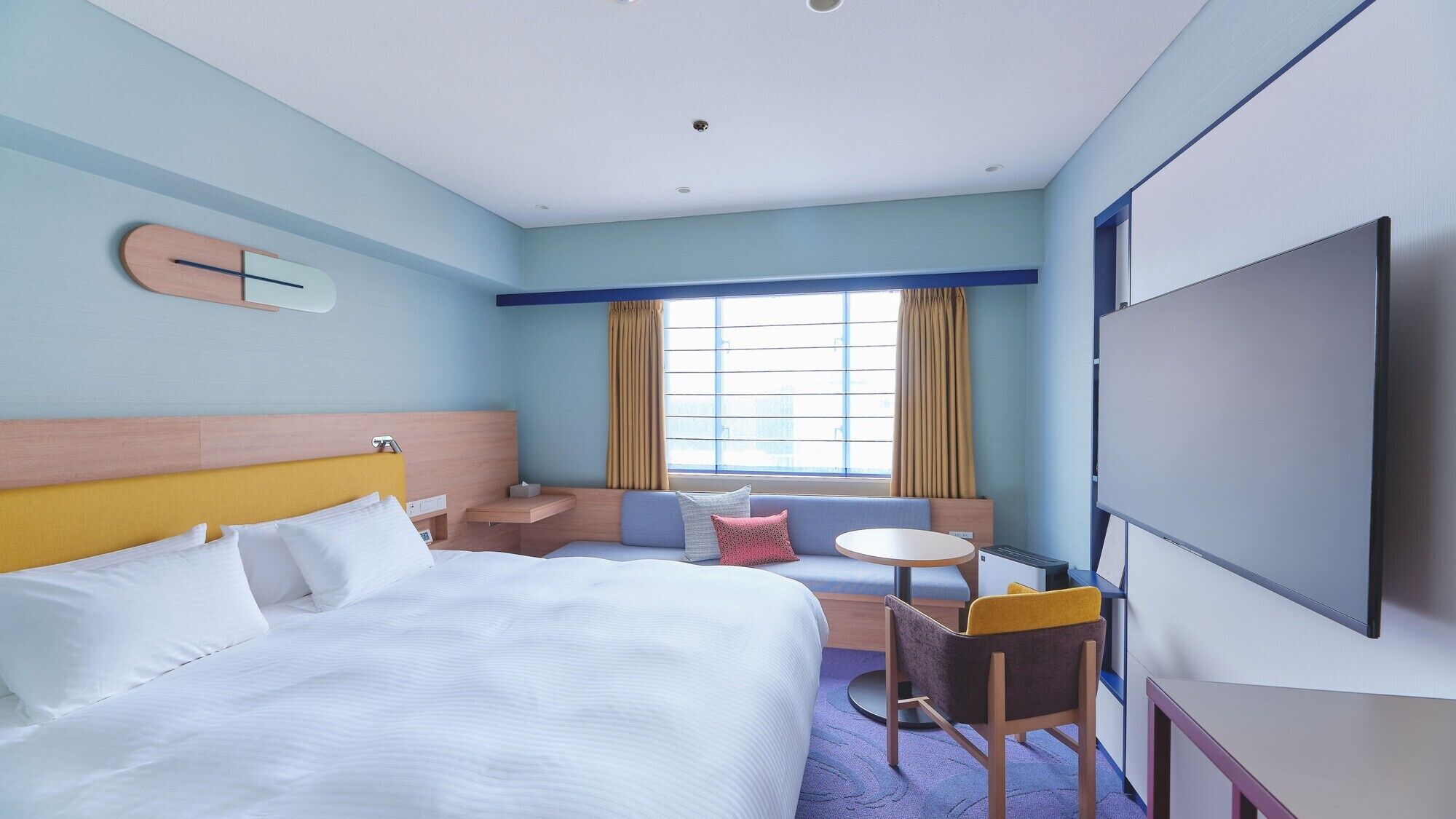 Quarto de hotel com cama, escrivaninha, cadeira, mesinha, televisão e janela com cortinas.