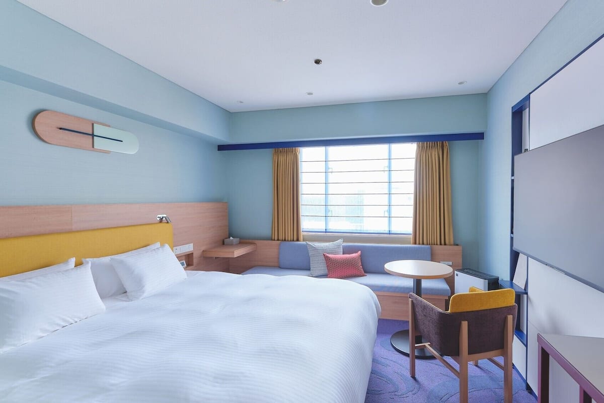 Une chambre d’hôtel comprenant un lit, un bureau, une chaise, une petite table, un téléviseur et une fenêtre avec des rideaux.