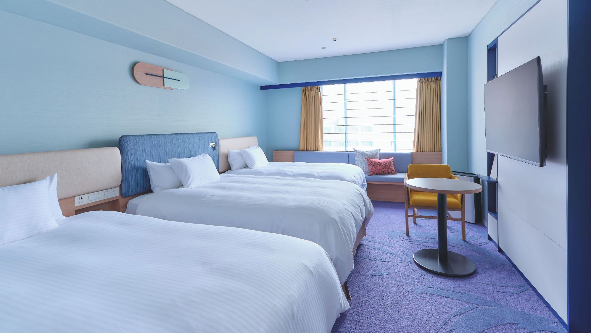 A hotel room with two beds, a small table, a chair, a TV, and a window with curtains.