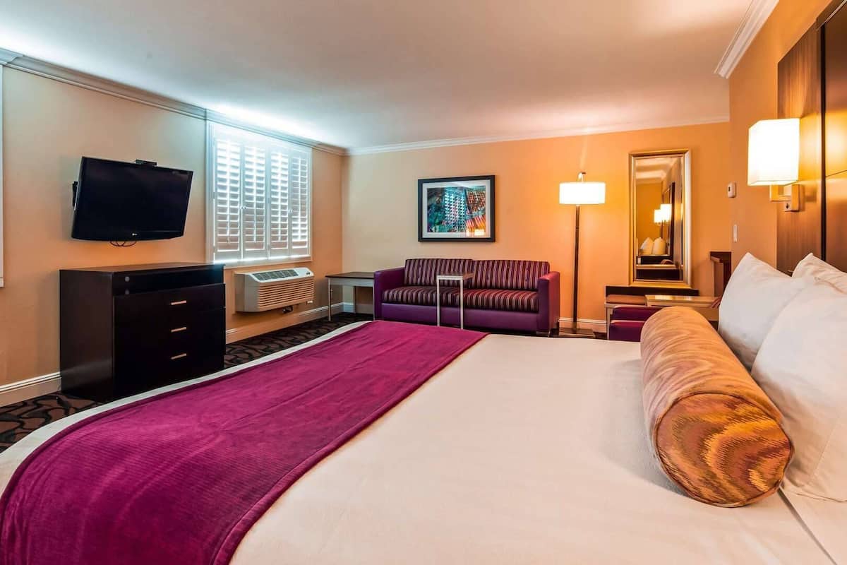 Een hotelkamer met een groot bed, een televisie, een bureau, een stoel en een raam met jaloezieën.