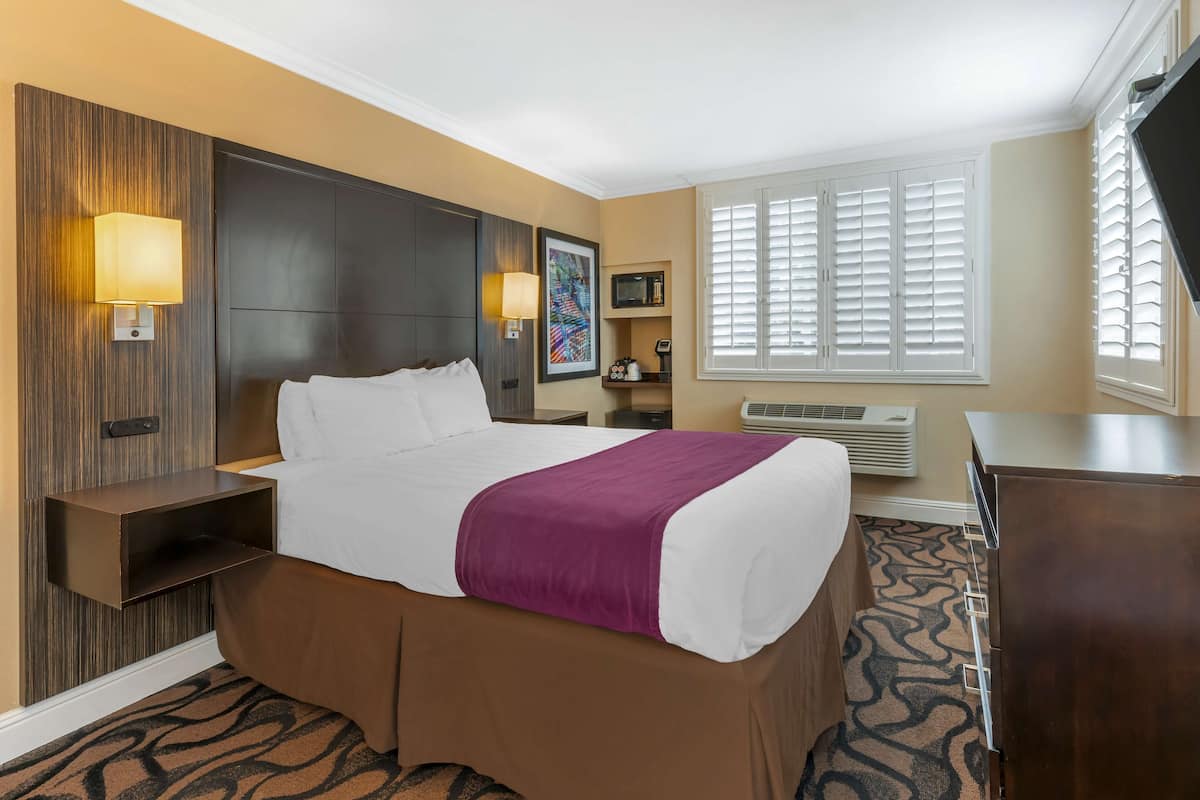 Hotelkamer met een groot bed, nachtkastje, bureau, stoel, televisie en raam met jaloezieën.
