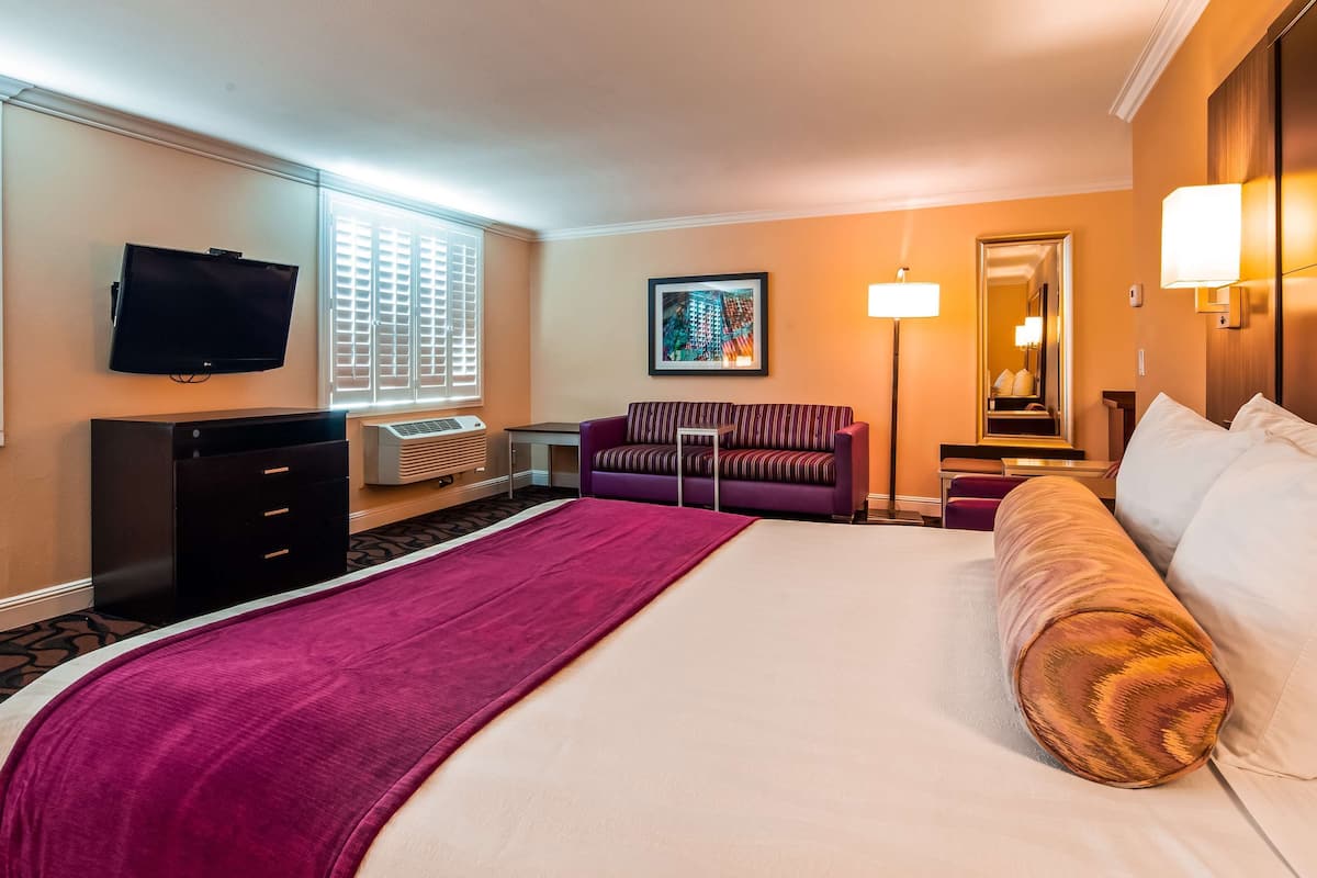 Een hotelkamer met een groot bed, een televisie, een bureau, een stoel en een raam met jaloezieën.
