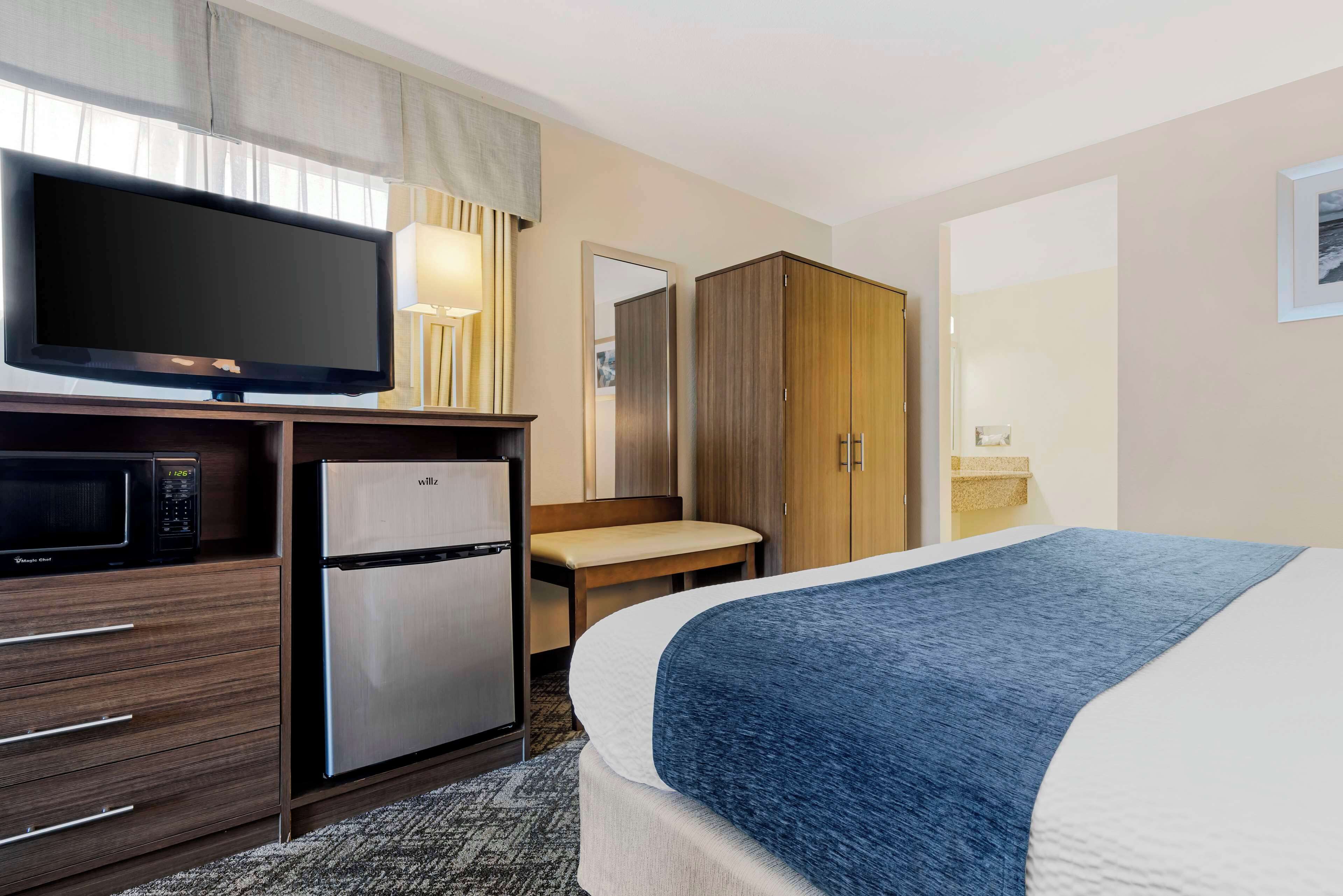 A hotel room with a bed, a television, a refrigerator, a microwave, a wooden wardrobe, a bench, and a mirror.