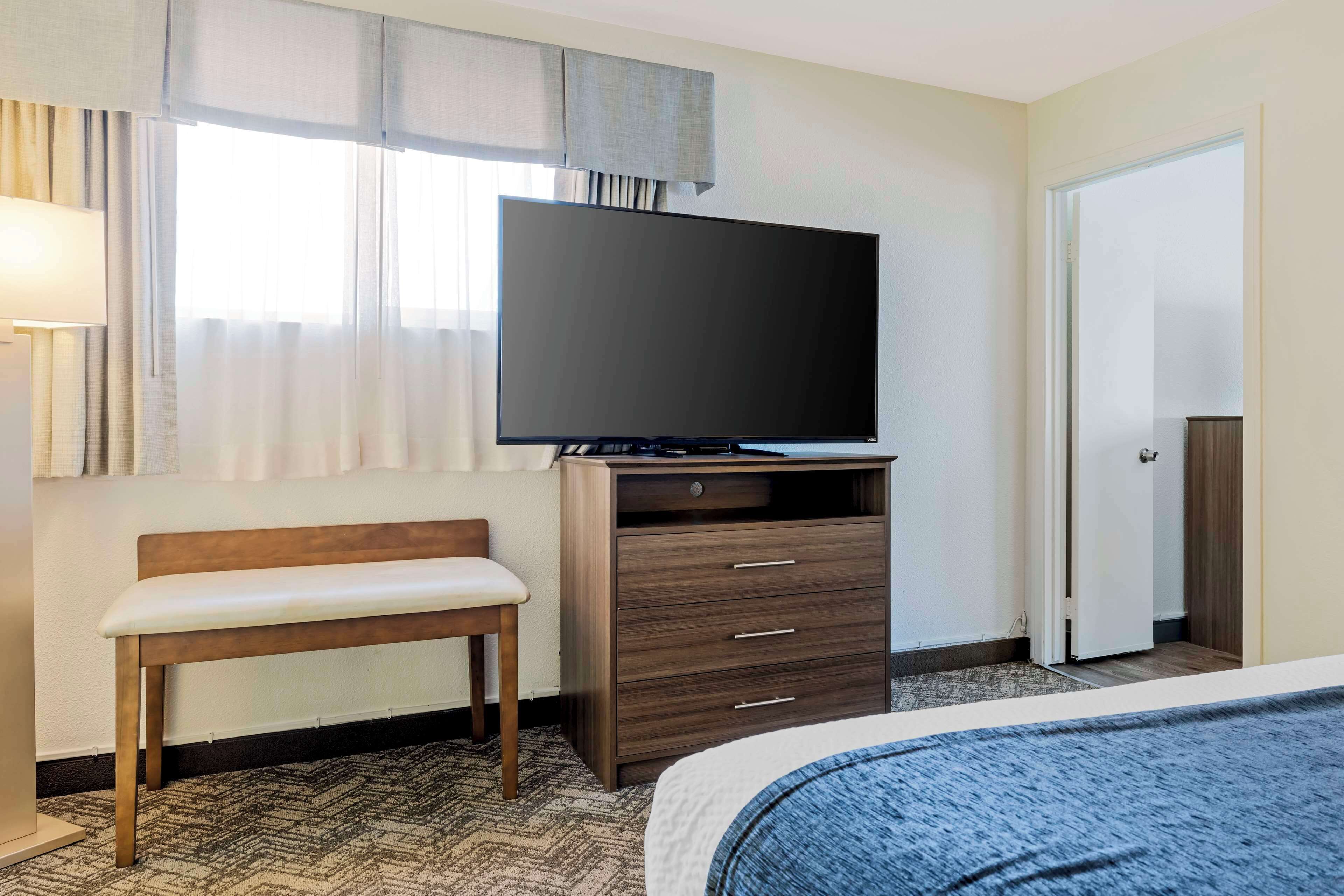 A hotel room with a bed, a wooden bench, a TV on a wooden stand, a door, and a window with curtains.