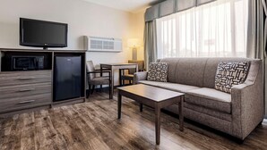 Habitación de hotel con televisión de pantalla plana, mini refrigerador, microondas, sofá con cojines estampados, mesa de centro, escritorio con lámpara y ventana grande con cortinas.