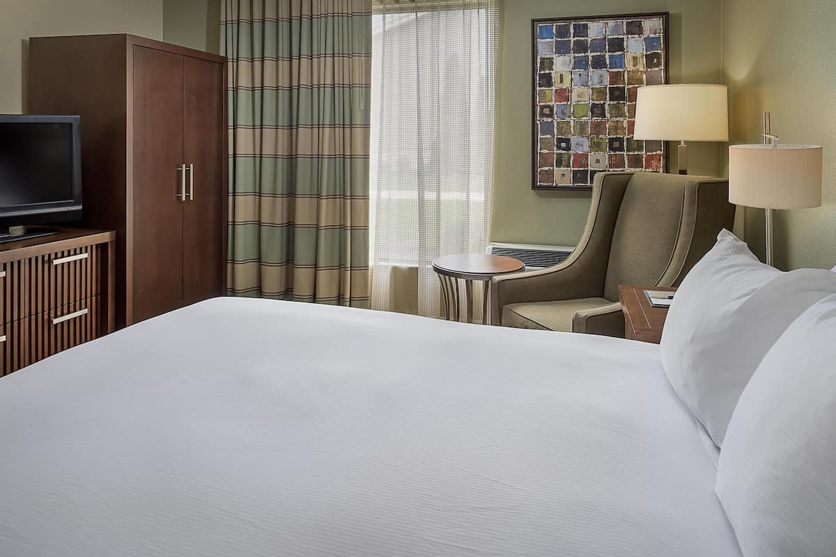 A hotel room with a bed, a television on a wooden cabinet, a chair, a small table, a lamp, and a curtain.