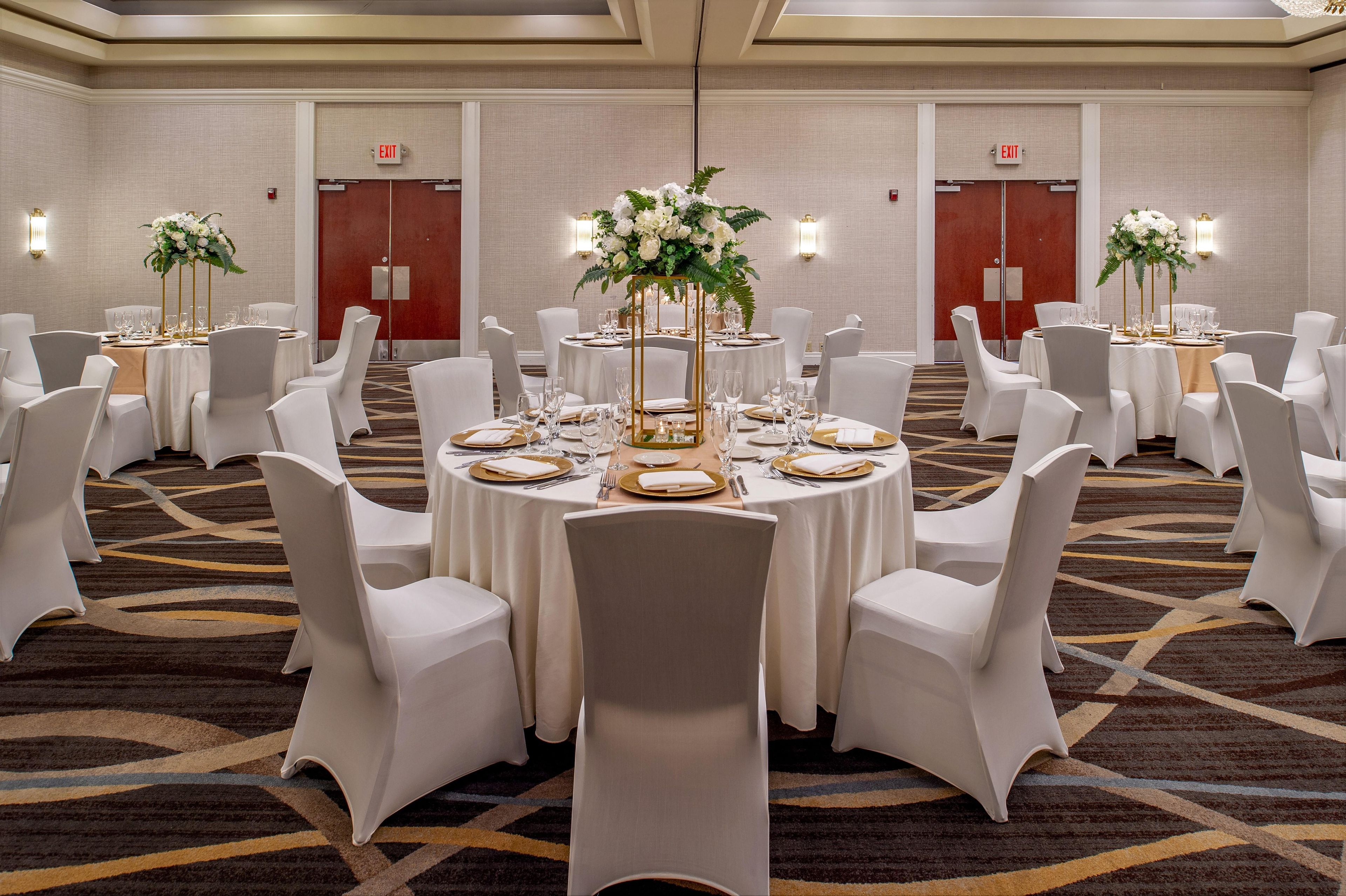 Un salón de banquetes con mesas redondas, sillas blancas y centros de mesa florales.