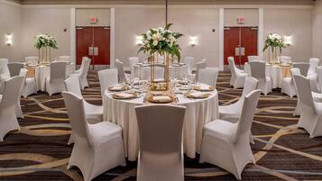Un salón de banquetes con mesas redondas, sillas blancas y centros de mesa florales.