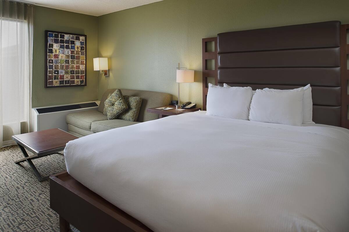 A hotel room with a large bed, a bedside table, a lamp, a small table, a sofa, and a wall art piece.