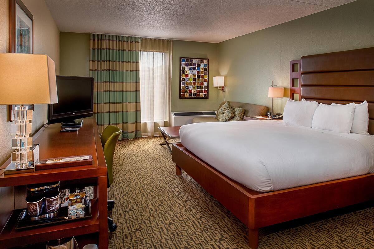 A hotel room with a large bed, a desk with a television, a chair, a small table, and a window with curtains.