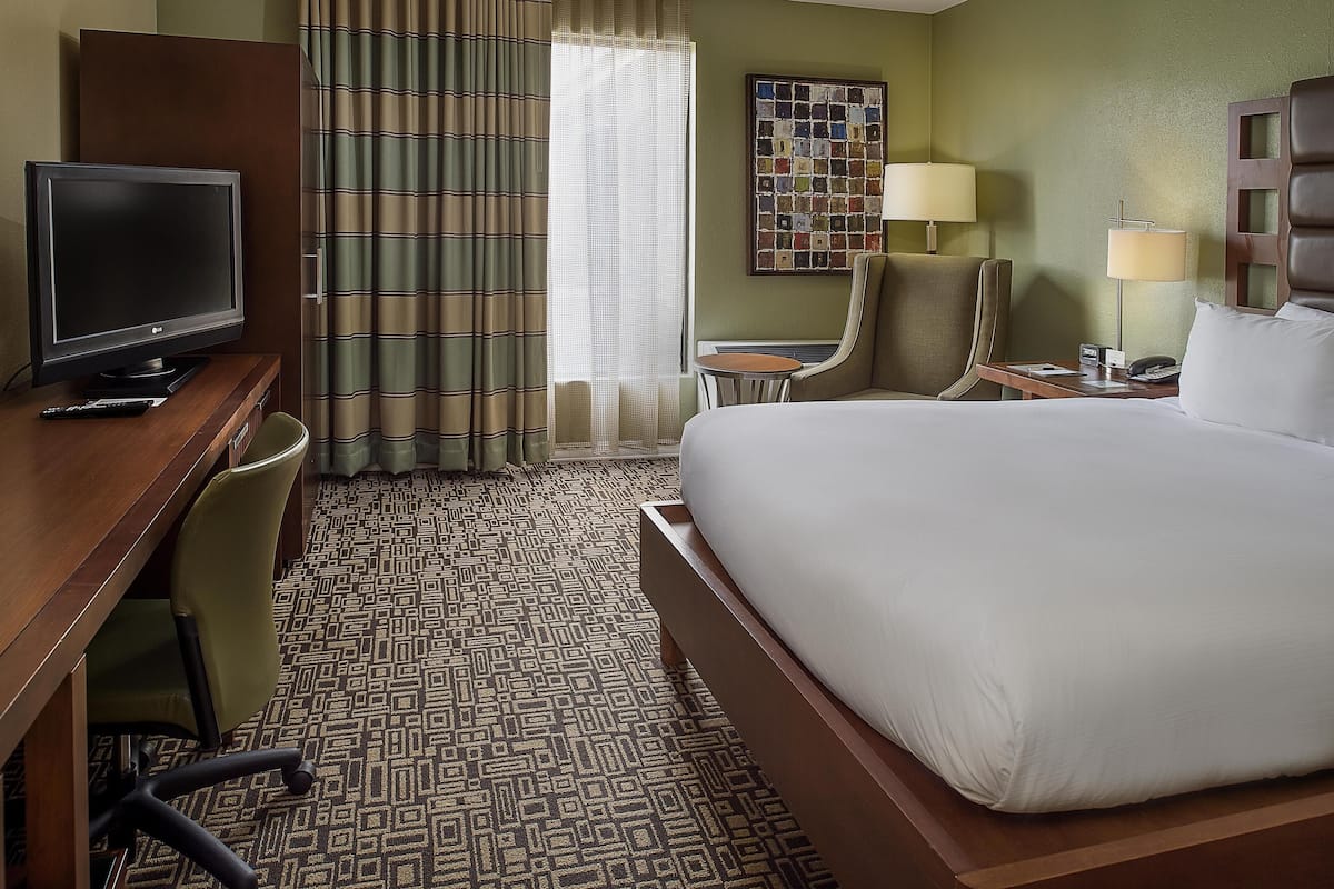A hotel room with a bed, a desk with a TV, a chair, a small table, and a lamp.