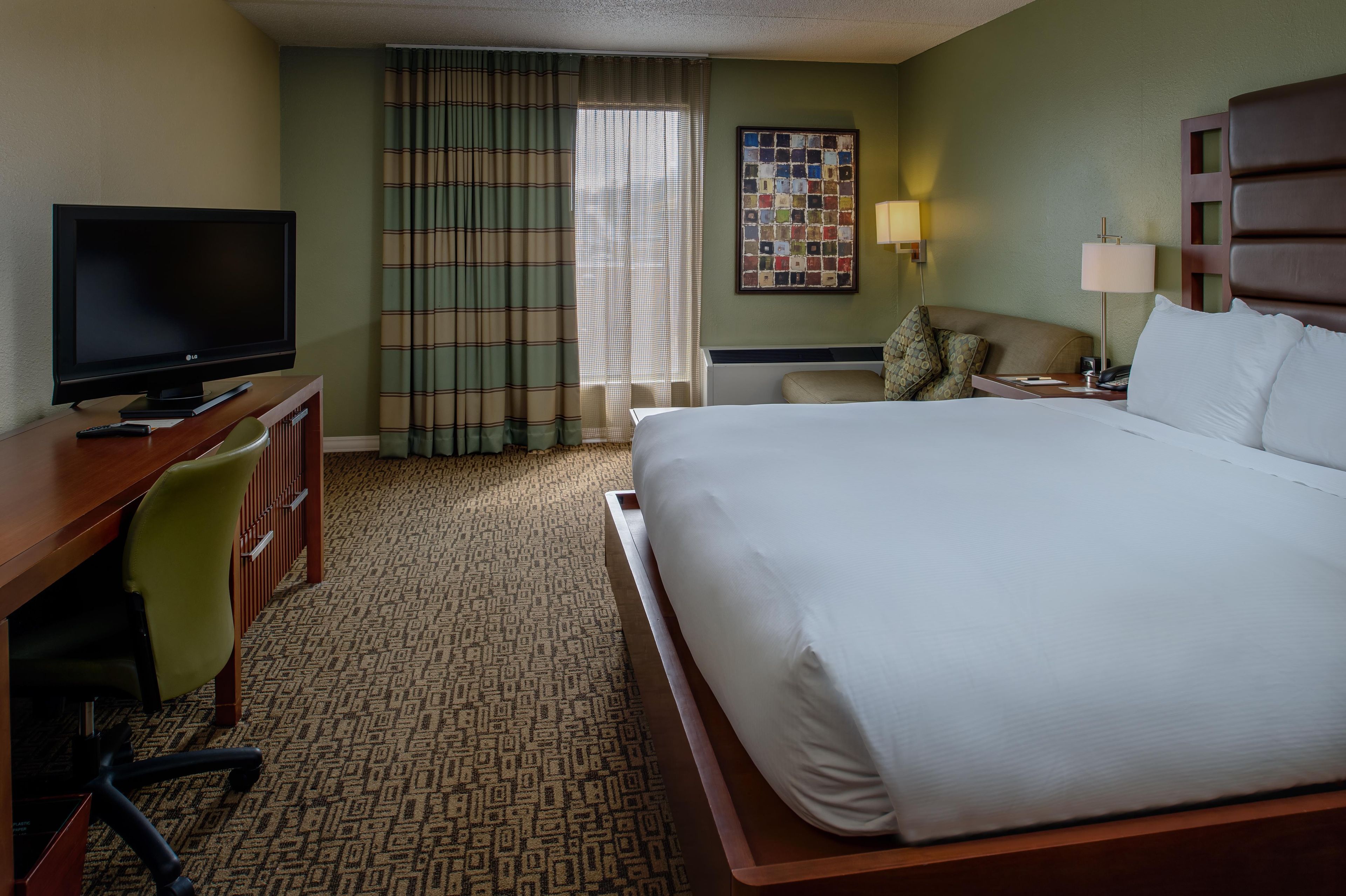 A hotel room with a bed, a desk with a television, a chair, a window with curtains, and a wall art piece.