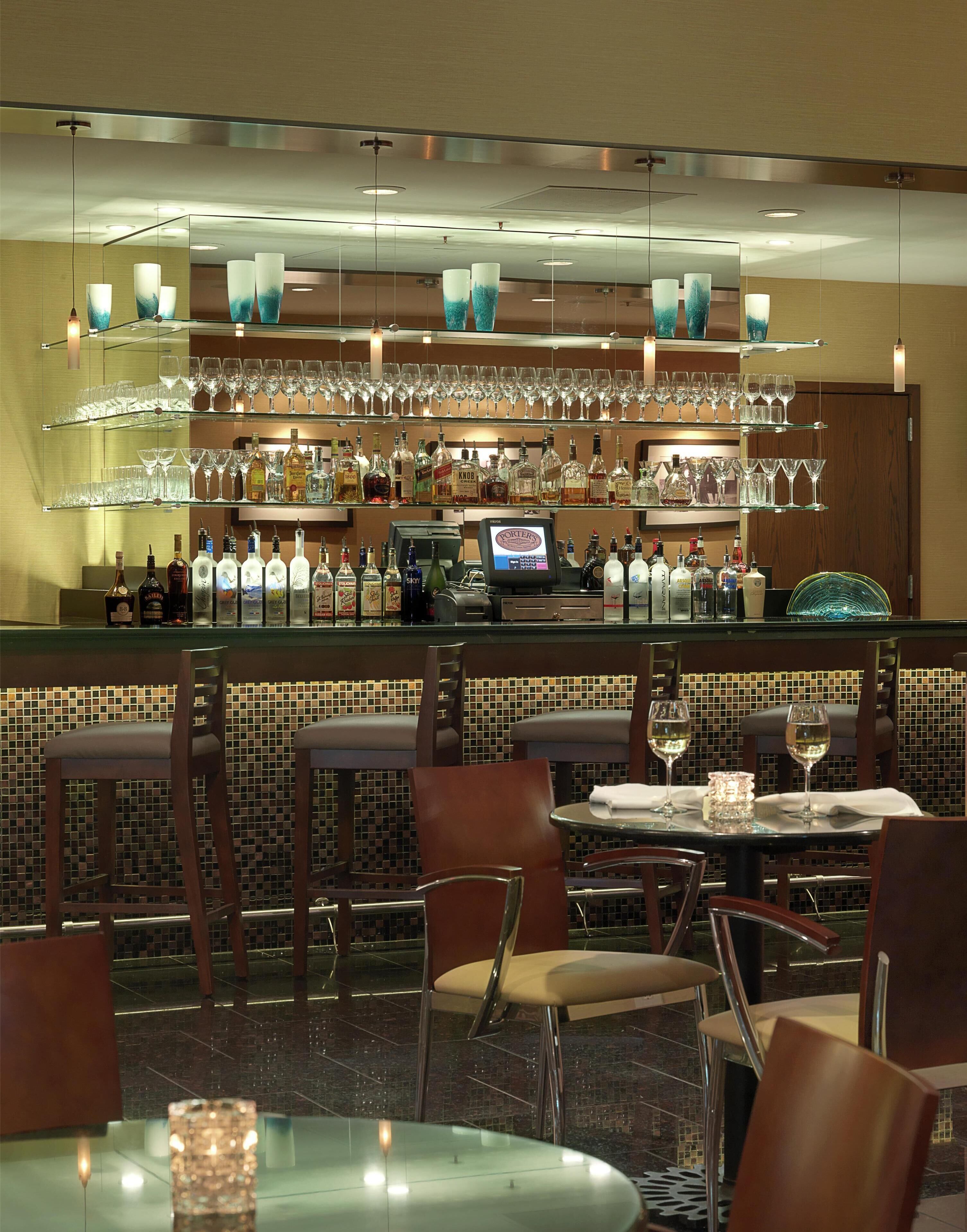 A bar area with multiple shelves of bottles and glasses, a cash register, and seating with tables and chairs.