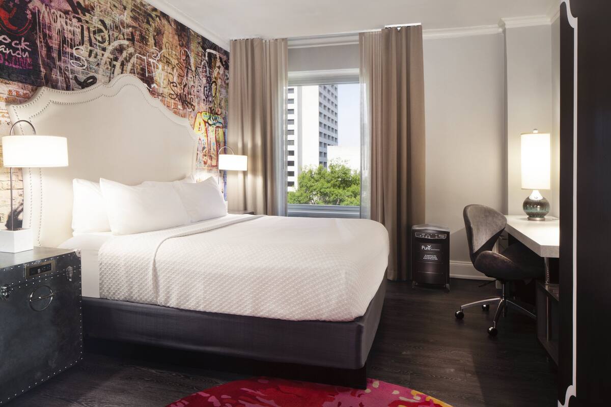 Habitación de hotel con una cama grande, un escritorio, una silla, una lámpara y vistas a un edificio desde la ventana.