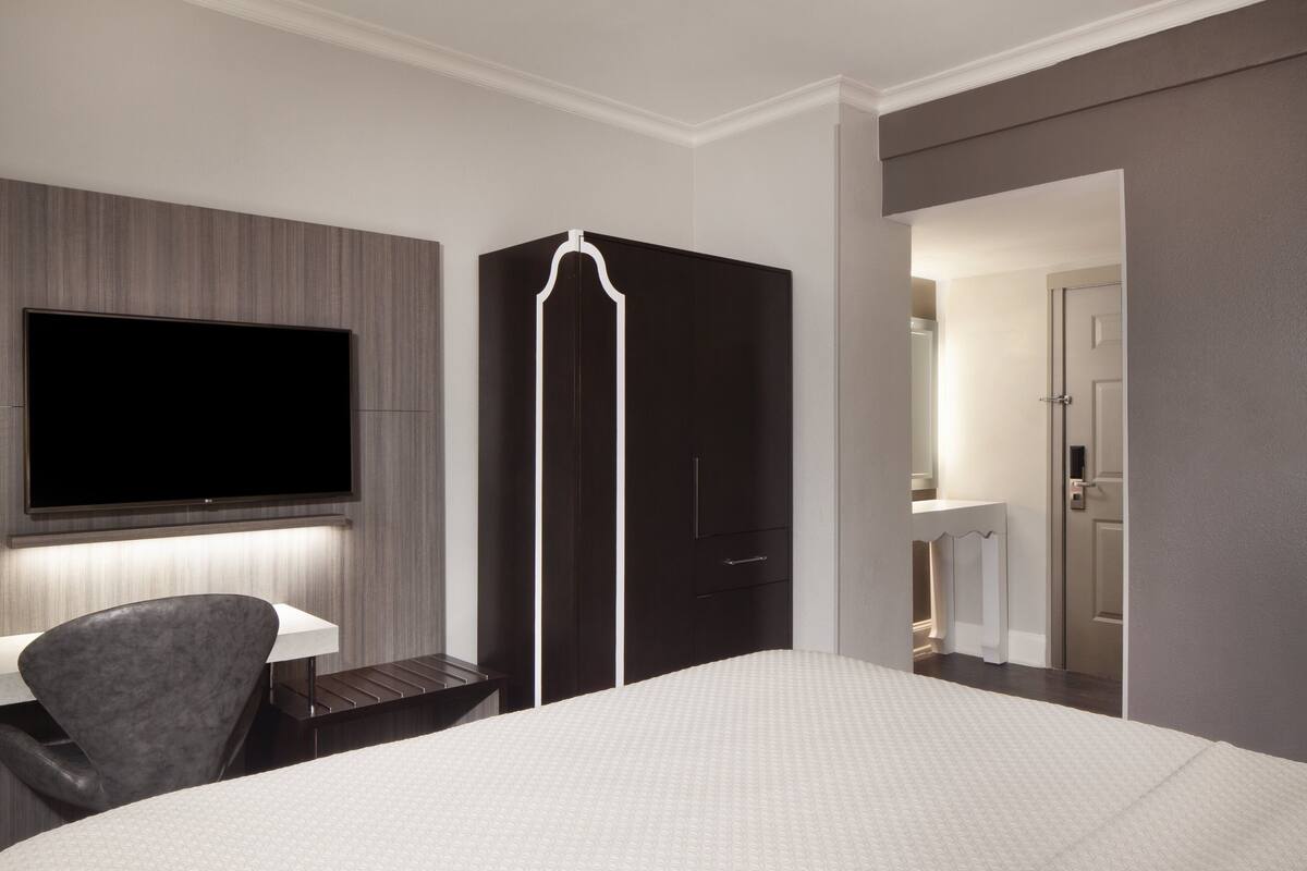 Una habitación de hotel con cama, escritorio con silla, televisor montado en la pared y armario.
