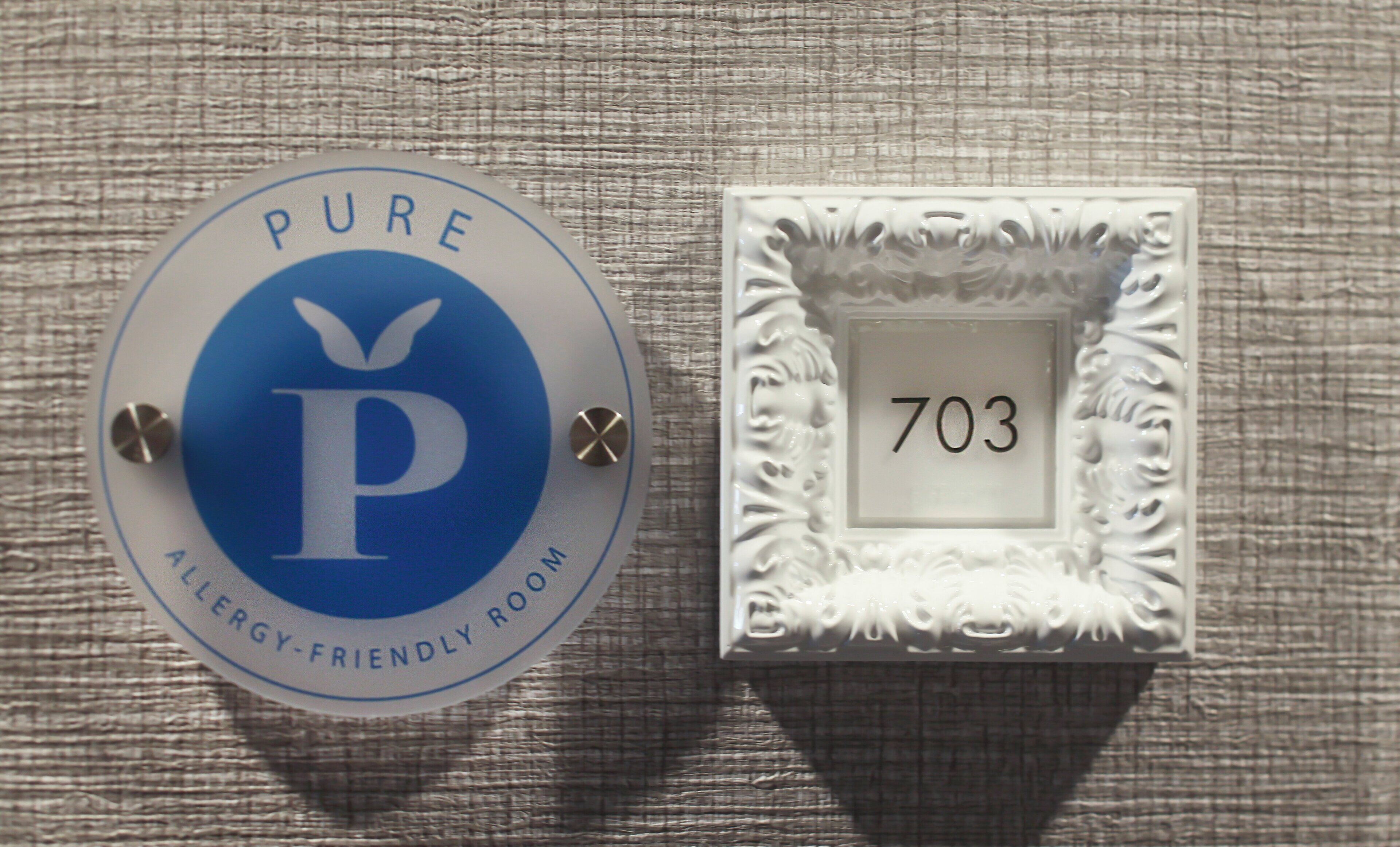 Un panneau circulaire portant les mentions « PURE » et « CHAMBRE ADAPTÉE AUX PERSONNES ATTEINTES D’ALLERGIES », et un panneau carré avec le numéro « 703 ».