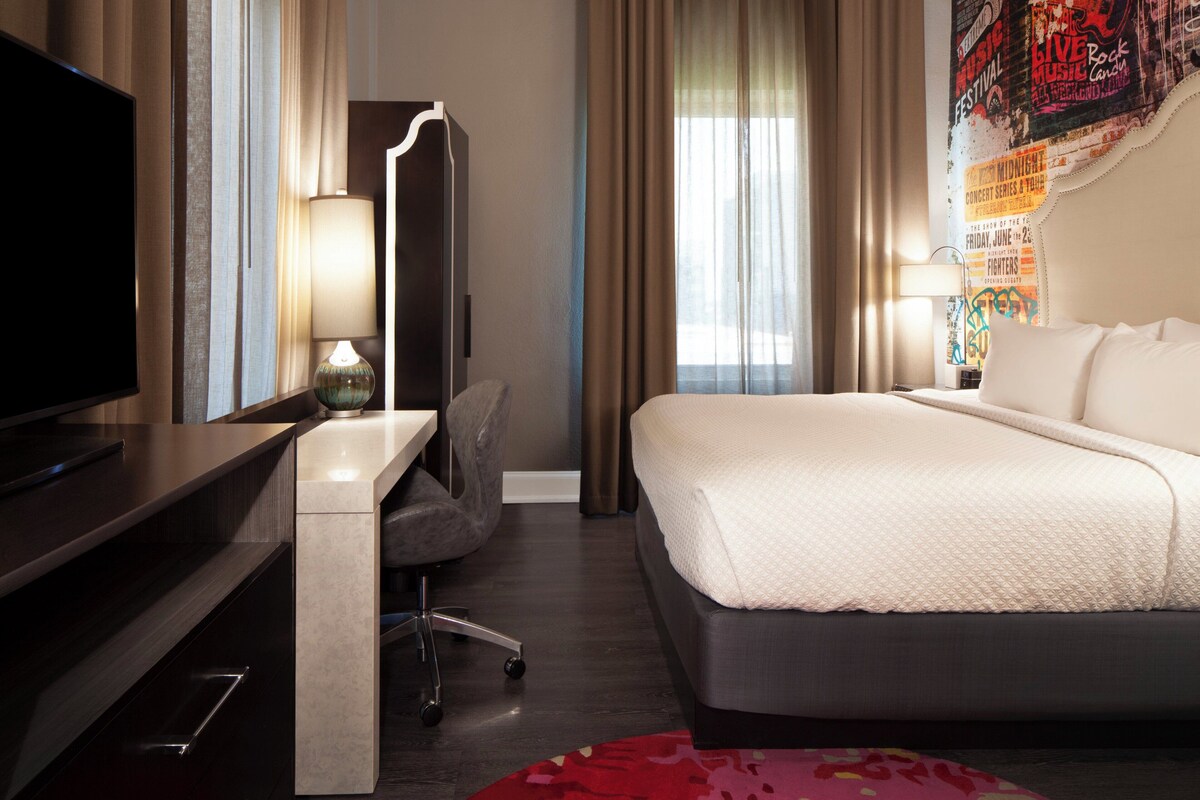 Una camera d'albergo con un letto, una scrivania con televisione, una sedia, una lampada e tende.