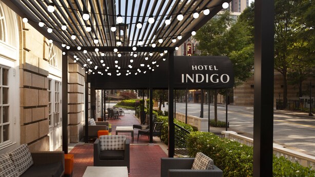 Un patio de hotel con mesas al aire libre, una zona cubierta con luces colgantes y un cartel que dice "HOTEL INDIGO".