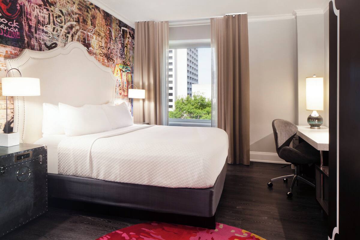 Habitación de hotel con una cama grande, un escritorio, una silla, una ventana con cortinas y un mural de pared colorido.