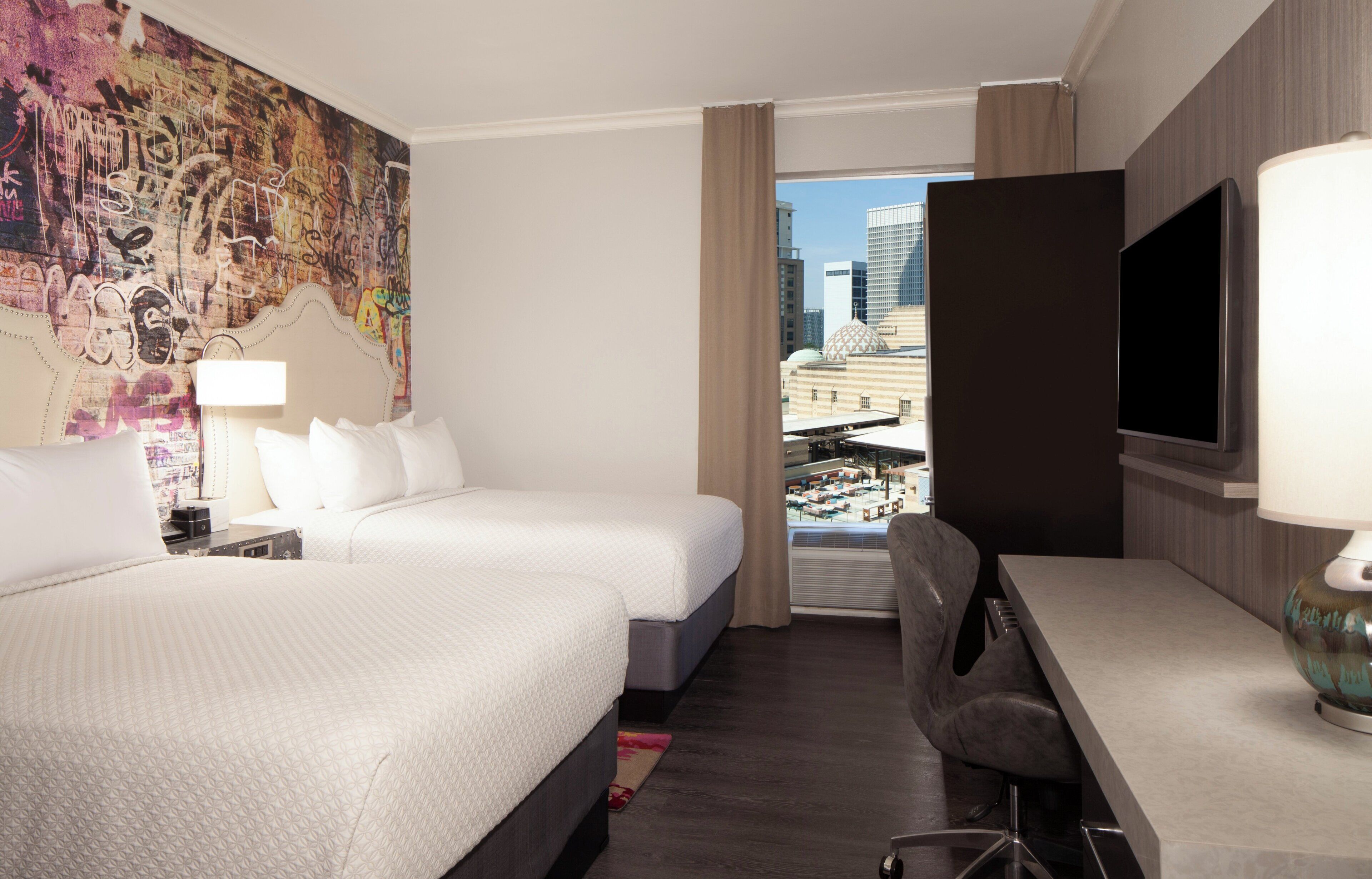Een hotelkamer met twee bedden, een bureau, een televisie en een groot raam met uitzicht op de stad.