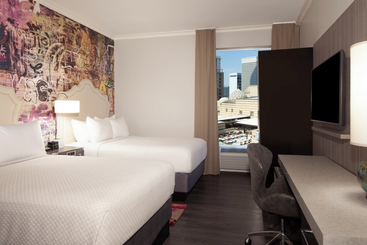 Camera d'hotel con due letti, una scrivania, una TV e un'ampia finestra vista città.